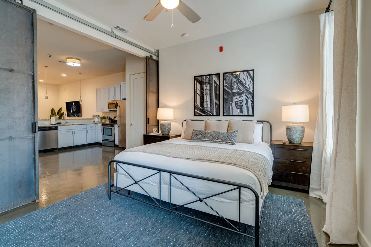 A stylish, sunlit bedroom with a plush bed adorned with decorative pillows, flanked by modern nightstands and warm lamps. Tasteful wall art adds character, while an inviting kitchen area is visible in the background, suggesting a seamless blend of relaxation and functionality.