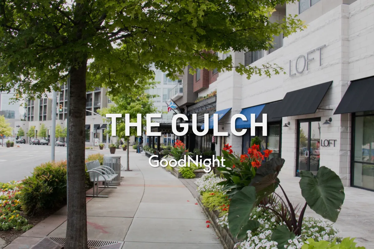 A lively streetscape with a tree-lined sidewalk, colorful flower beds, and charming storefronts. People are seen strolling and socializing in the bright outdoor setting, hinting at the vibrant local life and the appeal of neighborhood exploration.