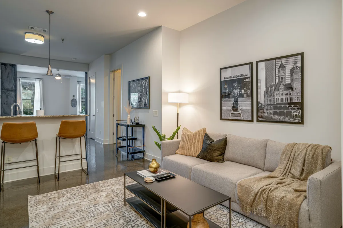 This image showcases a stylish, contemporary living area with a comfortable sofa, coffee table, bar stools at a kitchen counter, and modern artwork on the walls. The open layout seamlessly connects the living and kitchen spaces, complemented by warm lighting that enhances the cozy yet urban vibe.