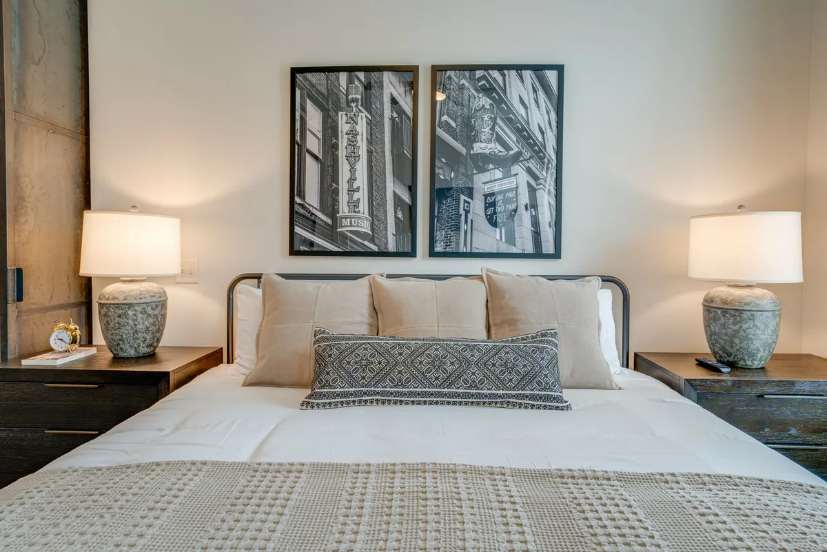 A tranquil bedroom highlighted by a king-sized bed, crisp white linens, framed artwork above the headboard, and matching nightstands with soft-glow lamps on either side. Gentle natural light filters in, complemented by blackout curtains for privacy and restful sleep.