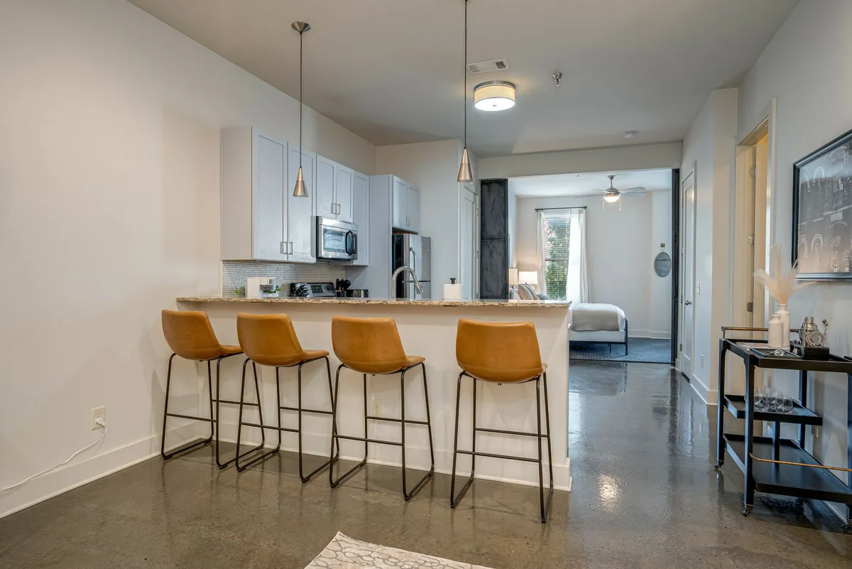 This image features a bright, modern kitchen with a central island, bar stools for casual dining, sleek stainless steel appliances, and ample natural light streaming in through a nearby window. The scene exudes a welcoming, functional atmosphere ideal for both cooking and socializing.