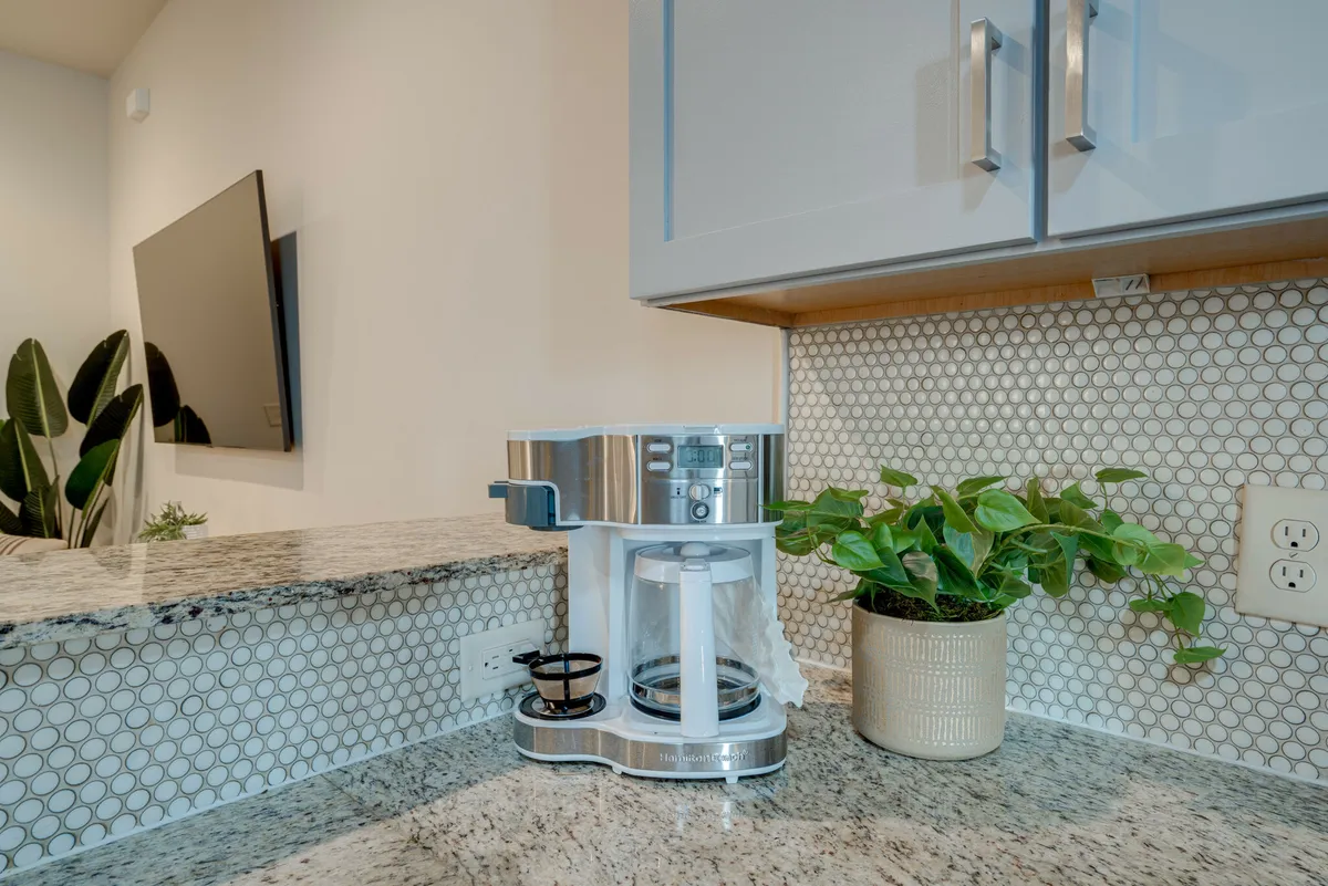The image shows a portion of the kitchen with granite countertops, a modern backsplash, a coffee maker set up for morning routines, and a touch of greenery for added warmth. The compact setup feels both functional and homey.