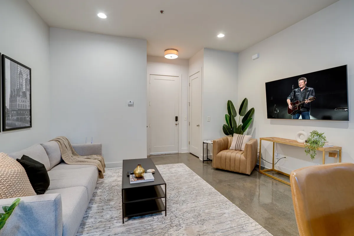 A cozy living room with a plush sofa, coffee table, large TV, and decorative greenery. The space is bathed in natural light from large windows, inviting relaxation, movie nights, or casual gatherings with friends and family.