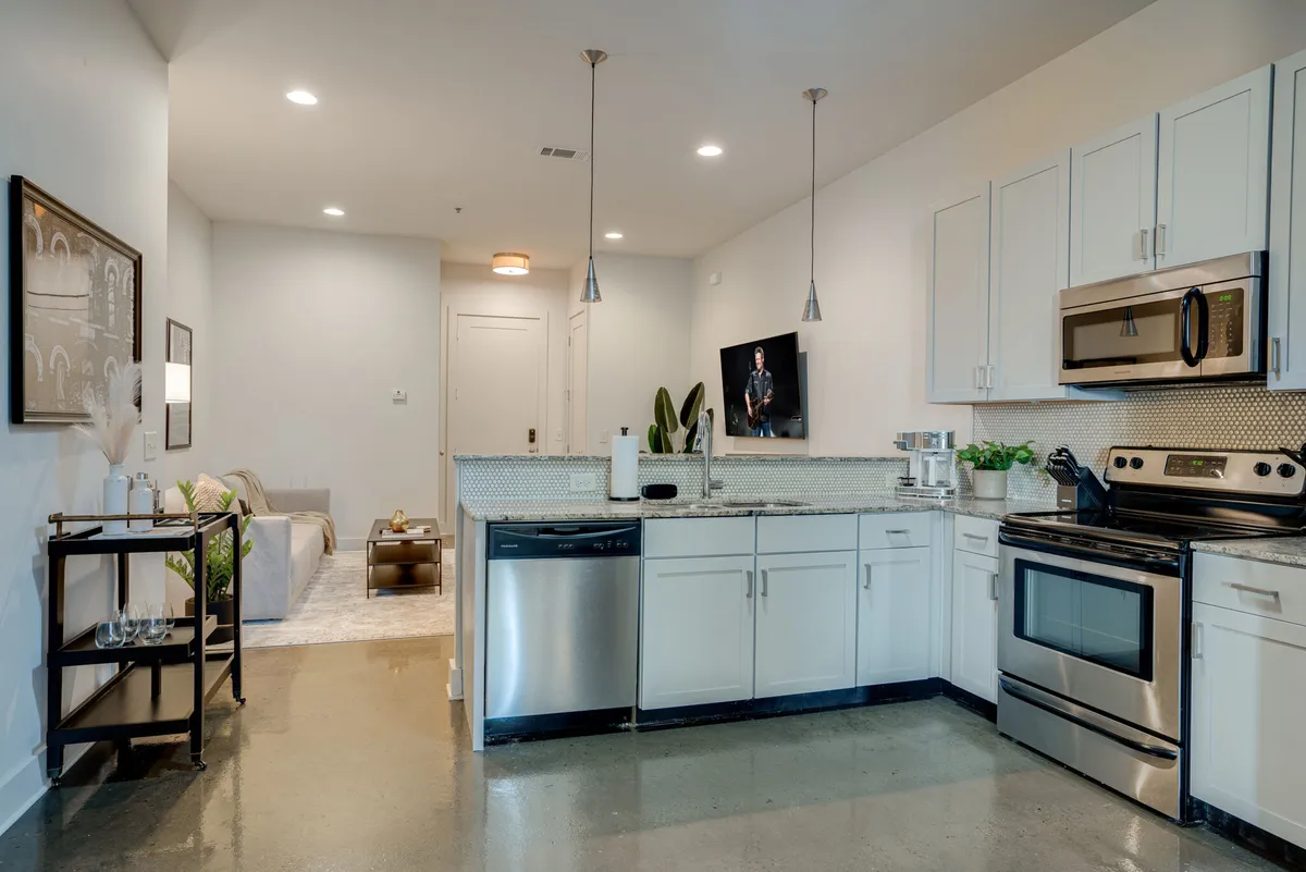 A welcoming, modern space blending the living room and kitchen. Visible are a comfortable sofa, stylish kitchen island with bar seating, stainless steel appliances, and eye-catching wall art, all under soft, inviting lighting.