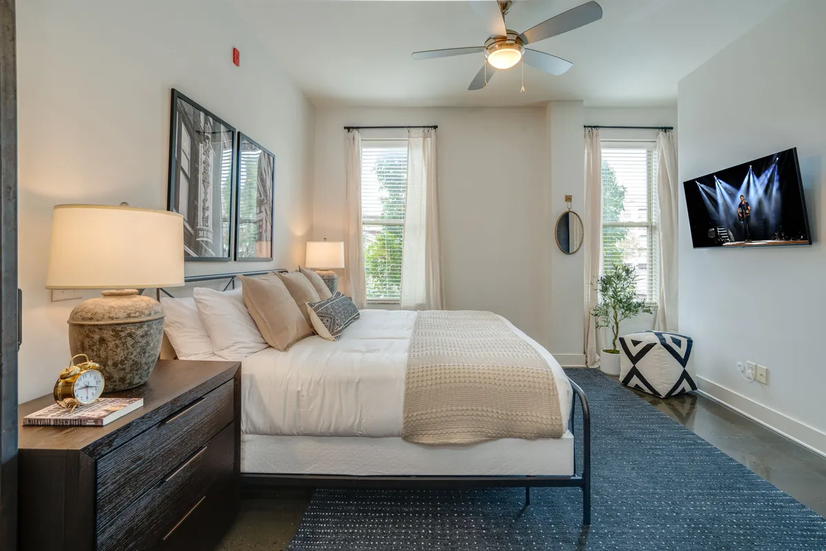 A stylish bedroom featuring a plush king-sized bed neatly made with designer linens, a nightstand with a lamp, a potted plant adding a touch of greenery, and a smart TV mounted opposite the bed. The overall feel is serene and inviting, ideal for relaxation and unwinding with entertainment.